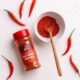 Close-up of cayenne powder in a ceramic bowl with spoon, fresh red chilies, and jar on white hex tile background.