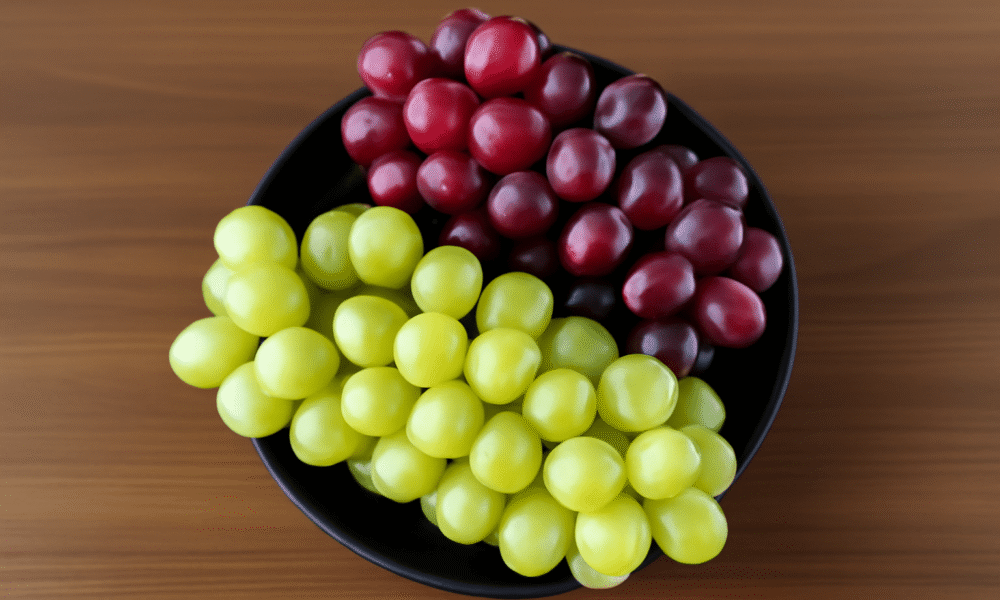 High-resolution digital photograph showcasing a black round ceramic bowl filled with clusters of white grapes and red grapes on a wooden surface.