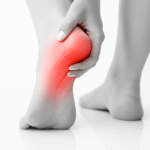 A black-and-white close-up photograph of a person's left foot showing heel pain. The heel area is highlighted in red to indicate inflammation or discomfort. The person's hand gently holds the heel, with the right foot blurred in the background. The scene is set on a reflective white surface.
