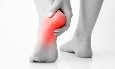 A black-and-white close-up photograph of a person's left foot showing heel pain. The heel area is highlighted in red to indicate inflammation or discomfort. The person's hand gently holds the heel, with the right foot blurred in the background. The scene is set on a reflective white surface.