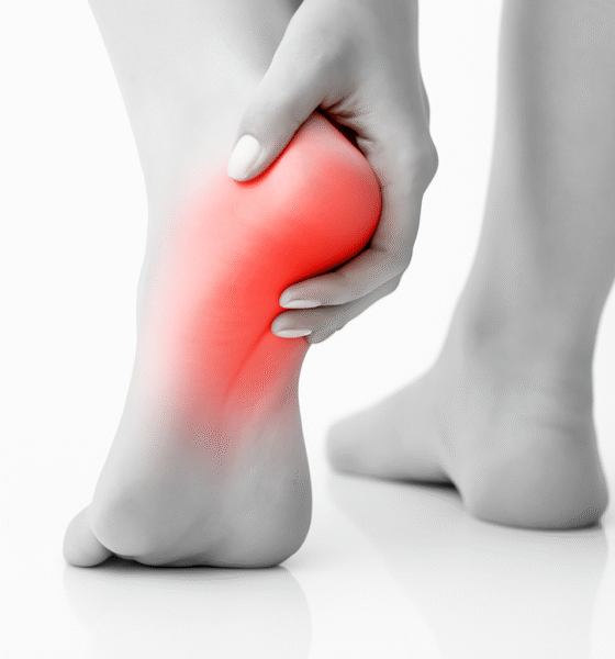 A black-and-white close-up photograph of a person's left foot showing heel pain. The heel area is highlighted in red to indicate inflammation or discomfort. The person's hand gently holds the heel, with the right foot blurred in the background. The scene is set on a reflective white surface.