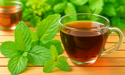 A close-up photo of a transparent glass cup filled with dark amber herbal tea, placed on a wooden surface. Fresh green mint leaves are arranged in the foreground, with blurred greenery and another copper cup visible in the background, all softly lit by natural light.