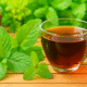 A close-up photo of a transparent glass cup filled with dark amber herbal tea, placed on a wooden surface. Fresh green mint leaves are arranged in the foreground, with blurred greenery and another copper cup visible in the background, all softly lit by natural light.