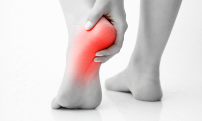 A black-and-white close-up photograph of a person's left foot showing heel pain. The heel area is highlighted in red to indicate inflammation or discomfort. The person's hand gently holds the heel, with the right foot blurred in the background. The scene is set on a reflective white surface.