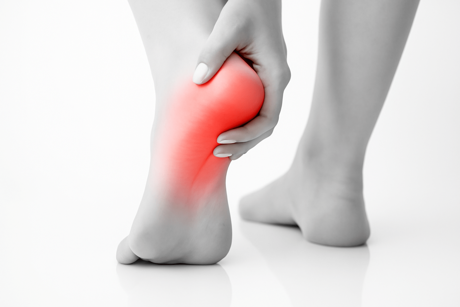 A black-and-white close-up photograph of a person's left foot showing heel pain. The heel area is highlighted in red to indicate inflammation or discomfort. The person's hand gently holds the heel, with the right foot blurred in the background. The scene is set on a reflective white surface.