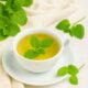 A cup of lemon balm tea with fresh leaves, set on a saucer, surrounded by green leaves and soft fabric in the background.