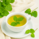 A cup of lemon balm tea with fresh leaves, set on a saucer, surrounded by green leaves and soft fabric in the background.