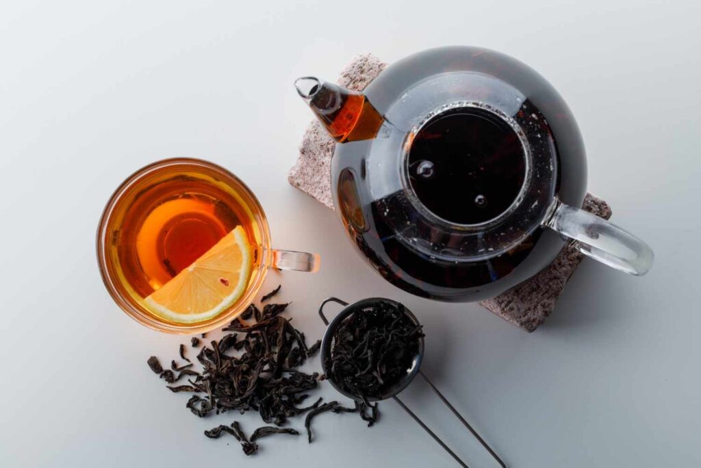 Glass teapot and cup filled with black tea and lemon slice, surrounded by loose black tea leaves on a white surface.