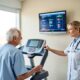An elderly male patient in a blue medical shirt stands on a treadmill while a female doctor monitors his cardiac stress test on the screen.