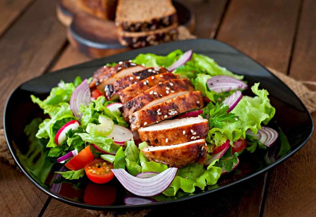 Grilled chicken breast served on a fresh vegetable salad with tomatoes, onions, and lettuce in a black plate for healthiest fast food meals.