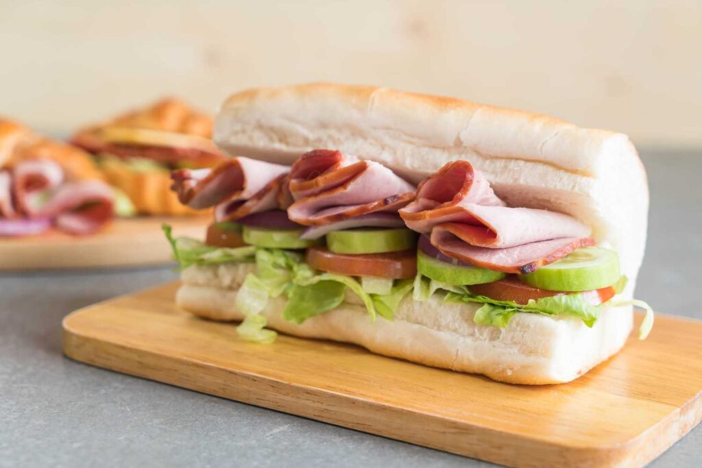 Sub sandwich with lean ham, fresh tomatoes, cucumbers, and lettuce served on a wooden plate for healthiest fast food meals.