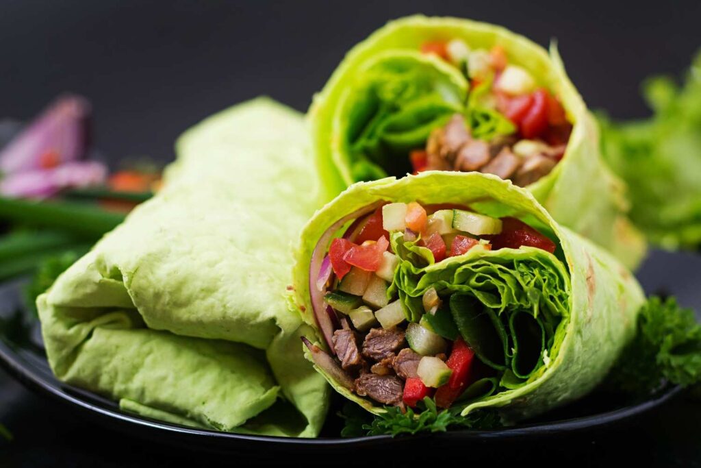 Veggie wraps with fresh lettuce, tomatoes, cucumbers, and beef filling, rolled and served on a black plate with healthiest fast food meals.