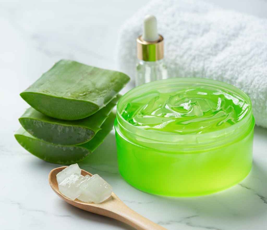 Aloe vera gel in a clear jar with fresh aloe leaf slices, dropper bottle, and towel on a marble countertop.